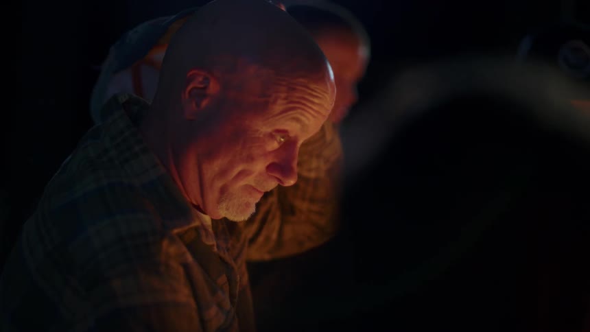 A man removes his baseball hat and scratches his hat as he sits in front of a fire outside