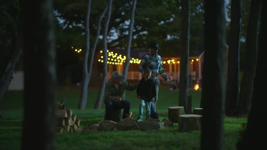 Boy and his grandfather sitting around an unlit campfire in a backyard while father walks over with wooden sticks
