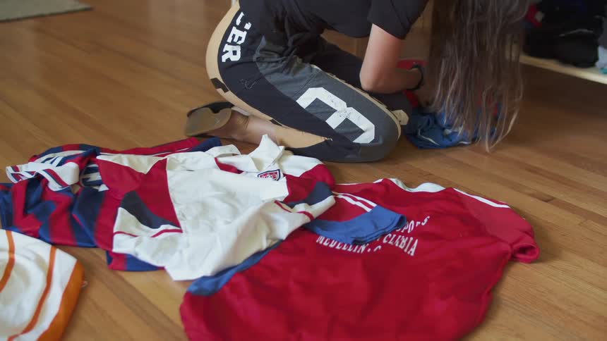 A woman kneeling down on a wooden floor inside of a room pulling different tops and jersey shirts out of a cabinet