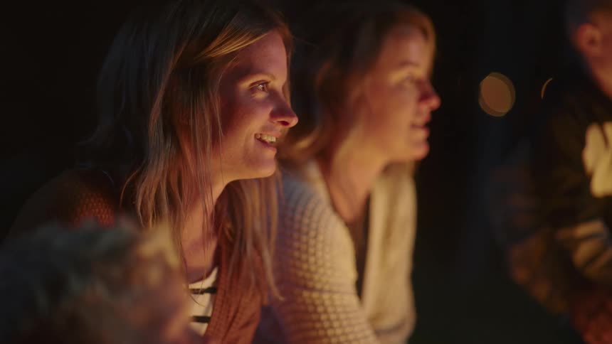 A woman smiles and laughs while sitting with friends around a campfire