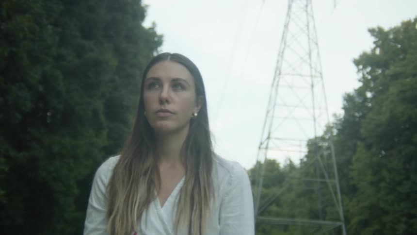 The camera shakily pans around a woman standing in a clearing in a heavily forested area