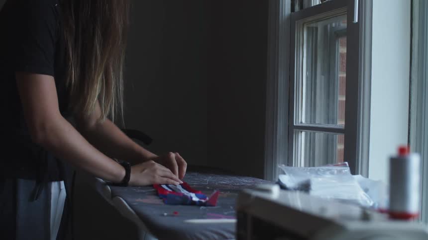 A woman stands in front of a window and sews white strips onto a blue and red piece of fabric