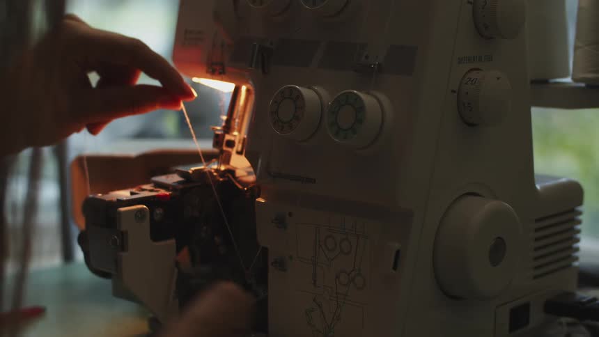 A woman sits in front of a sewing machine adjusting settings and pulling a piece of thread