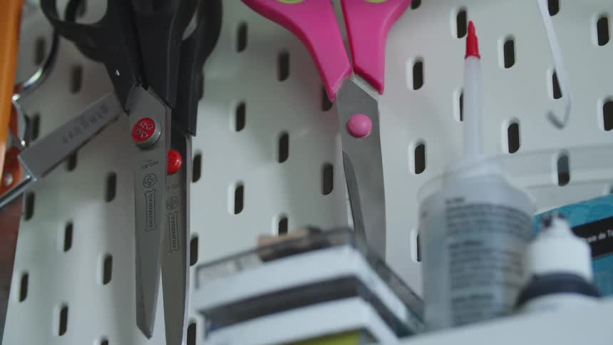 Pink and black scissors hanging from a white shelf next to other supplies