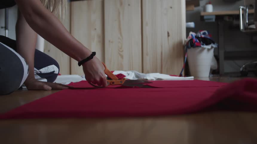 A woman is kneeling on the floor and cutting through a piece of red fabric with a pair of scissors