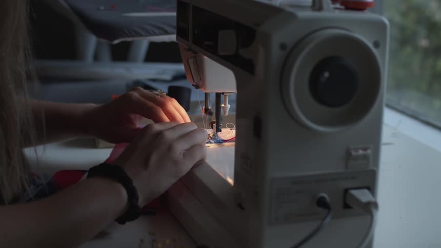 A woman pushed fabric through a sewing machine then cuts the thread with scissors