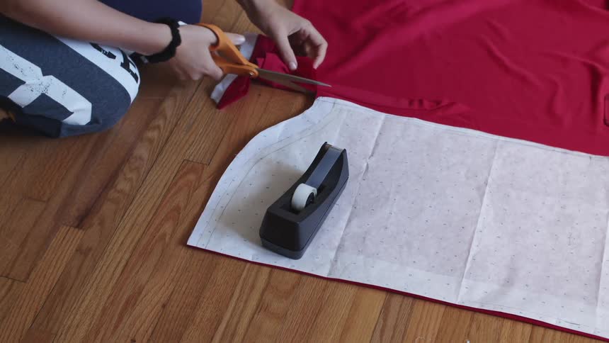 A woman sits on a hardwood floor cutting red fabric according to the white pattern