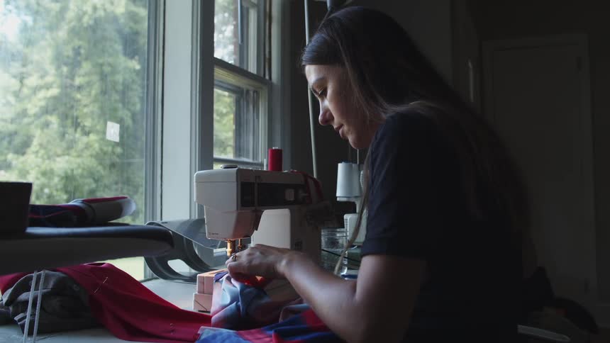 A woman sits at a sewing machine in front of a large window sewing fabric