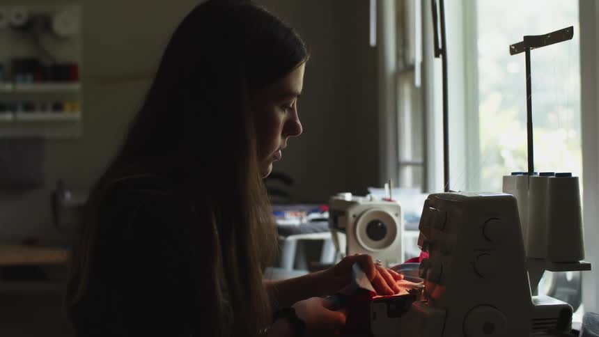 Woman inside of a room sitting in front of a sewing machine sewing a piece of fabric on it