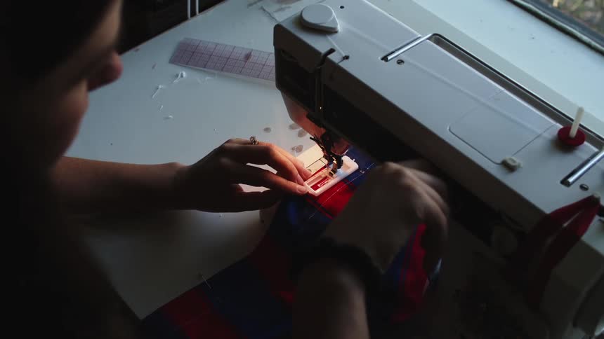 Woman finishes up on sewing a blue and red shirt under a sewing machine at a white table indoors