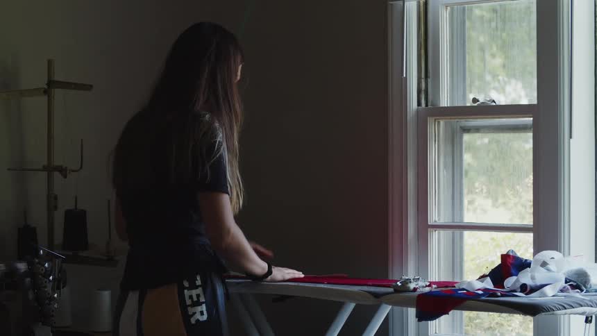 A woman stands at an ironing board in front of a window ironing clothes