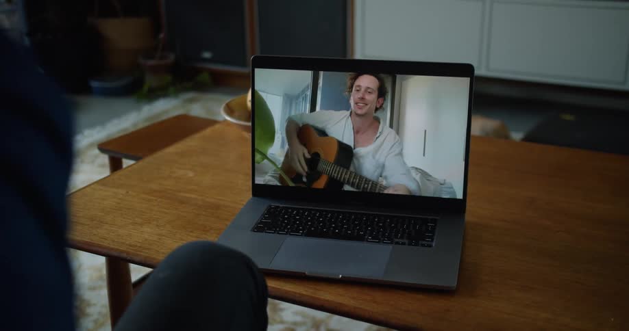 A laptop computer on a table in a living being used to web chat with man holding an acoustic guitar