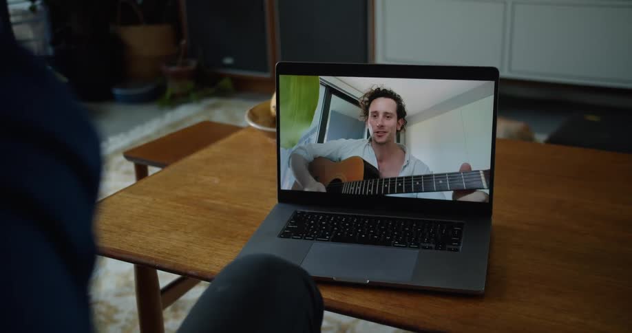 Man on a laptop computer screen in a living room holding an acoustic guitar and talking