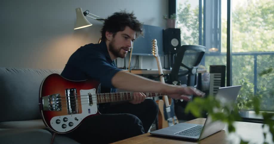 Man holding a white and brown electric guitar sitting on a couch in a living room waving goodbye to a webcam