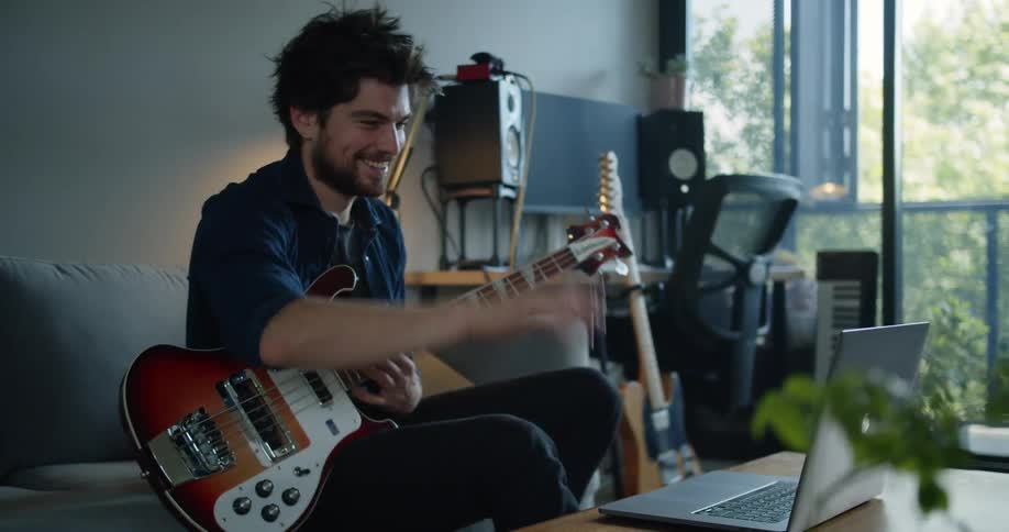 Man sitting down on a couch in his living room holding a guitar and talking on a laptop