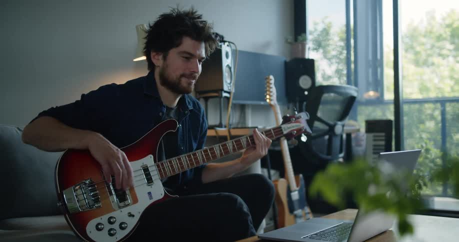 A smiling bearded man playing a guitar on a couch in a room in front of a laptop