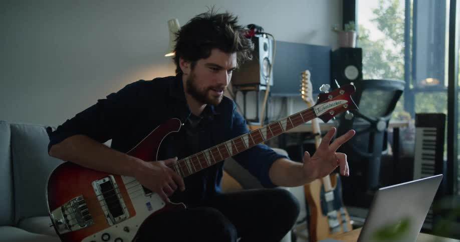 A man sits on a couch typing on a laptop while holding a guitar