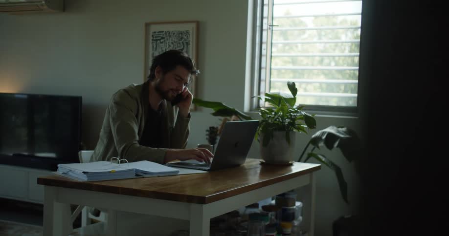 A man talks on the phone while typing on his laptop while working from home