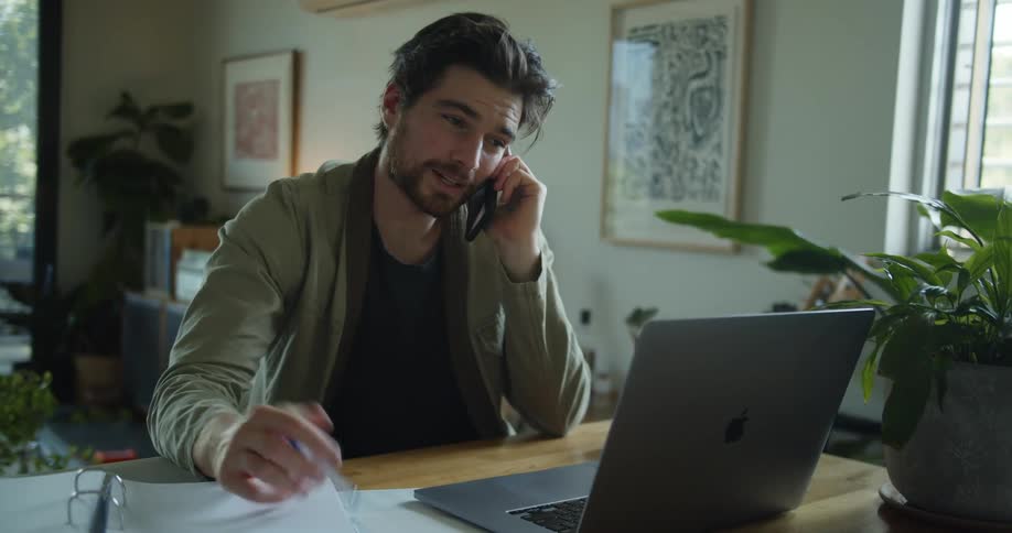 A man talks on his smartphone while sitting in front of a laptop at a desk in a house during the day