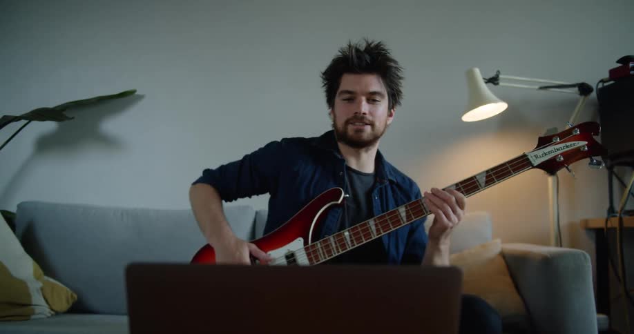 Man holding a bass guitar waves at a laptop web cam and gets read to play