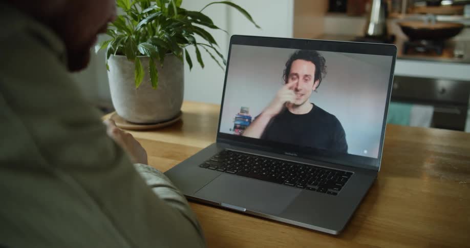 A person sits at a table drinking from a cup communicating with a man on a laptop