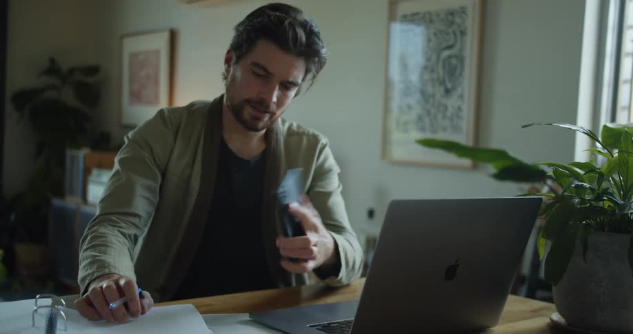 Young man working on a laptop from home hangs up a call on his smartphone
