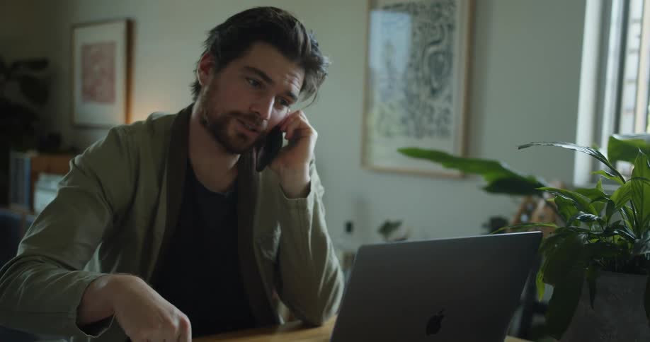 Man talking on a cellphone sitting in front of a laptop in a living room at a table