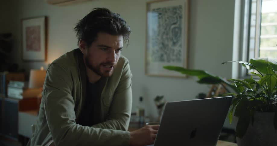 Man with a beard and a green jacket sits at table and makes a video call while drinking a cup of coffee