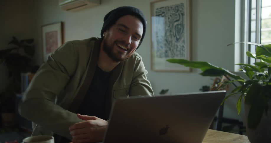 A man with a hat sitting on a chair by a desk talking on a laptop