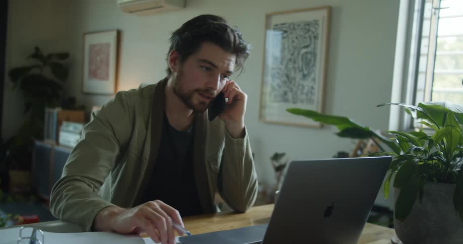 Man on a smartphone sitting at desk with paper work in front of him and a laptop computer