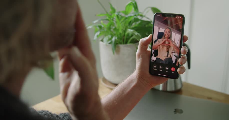 A man is facetiming with a woman throwing kisses back and forth while sitting at a desk indoors during the day