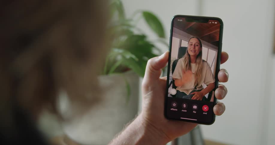 A man holds a phone displaying a woman throwing a kiss to him sitting indoors