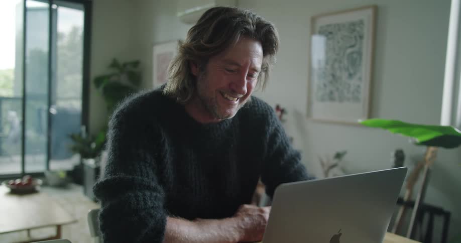 Man in a living room sitting at a table in front of an open silver laptop smiling and talking to a webcam