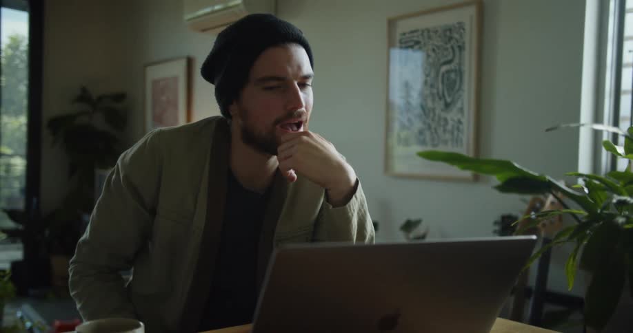 Man wearing a beanie talks to someone in a video call on a laptop at home