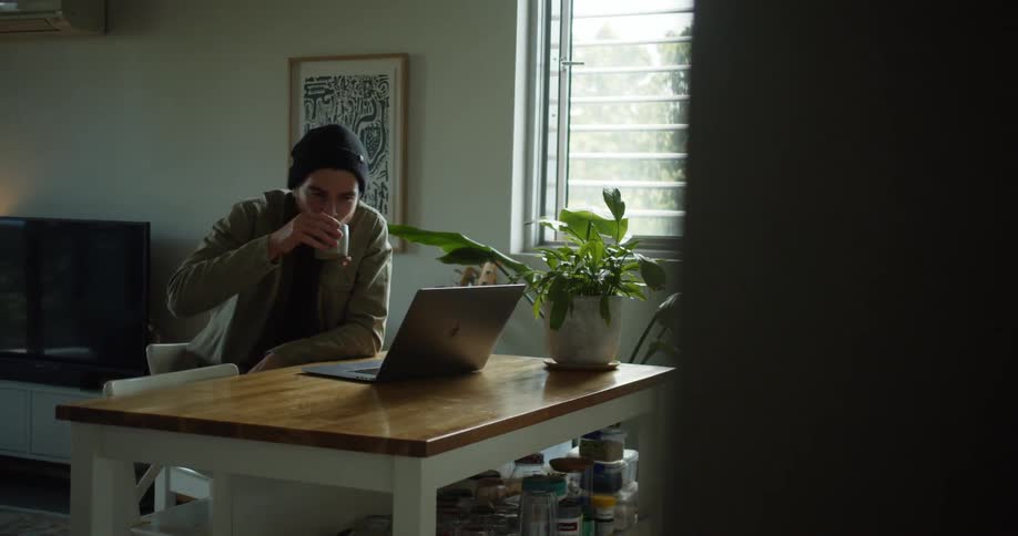 Man sitting at a small desk in a living room web chatting on a laptop and drinking from a mug during the day