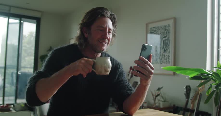 Man with long hair and a black pullover makes a video call on his smartphone while drinking a cup of coffee