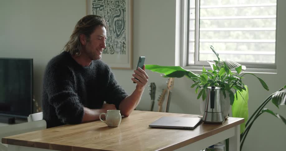 Man with long hair and a black sweater makes a video call on iphone 12 in the living room at home