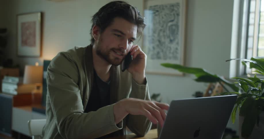 Man with a beard talks on the smartphone while using the laptop in the living room at home