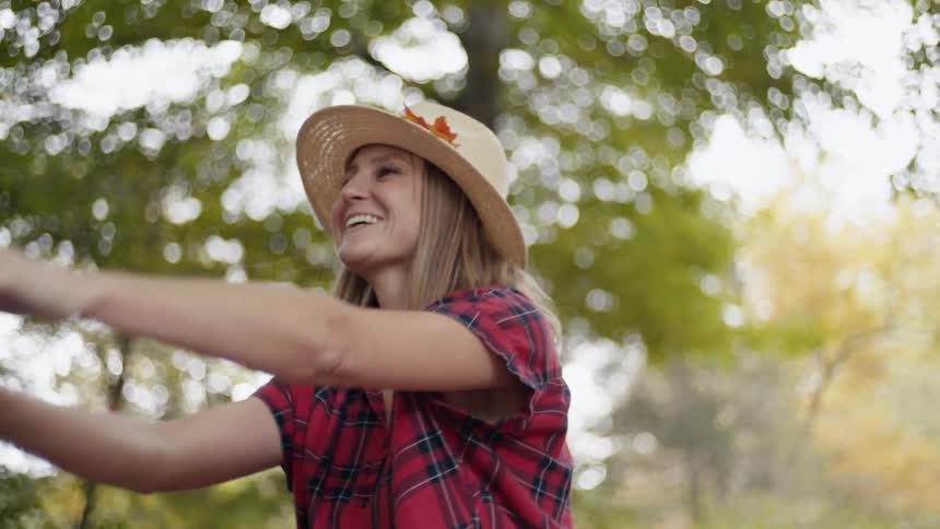 A woman wearing a hat and a red plaid shirt pushes a child in a swing outdoors with trees in the background