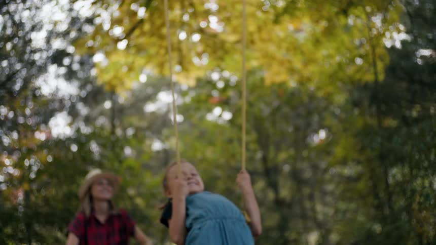 A mom pushes a swing that her daughter is standing on in the middle of trees