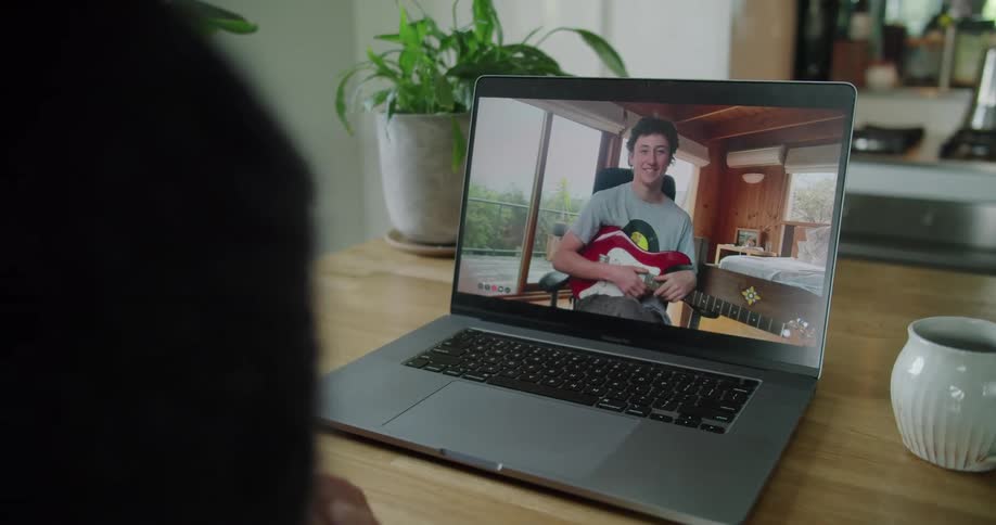 Man on a laptop computer screen sitting in a bedroom holding an electric guitar and talking