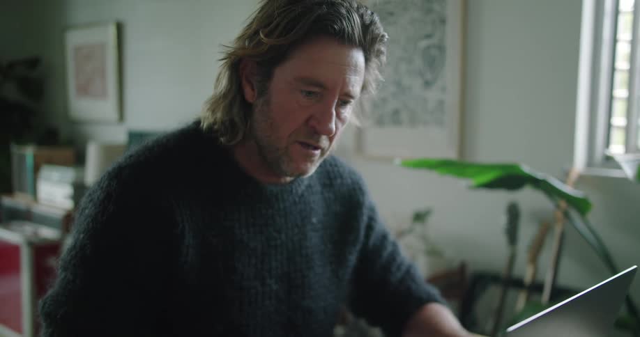 A man sits at a table at home typing on a laptop during the day