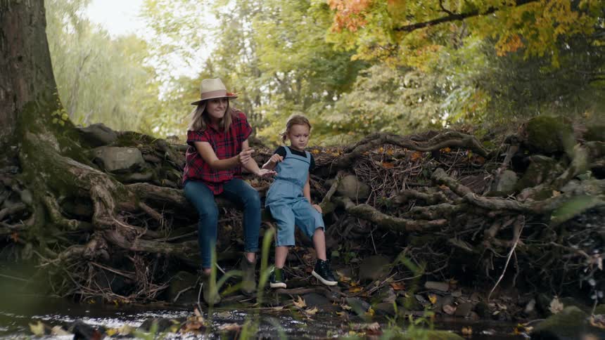Mother and daughter sitting on a large tree root next to a stream skipping rocks together at daytime