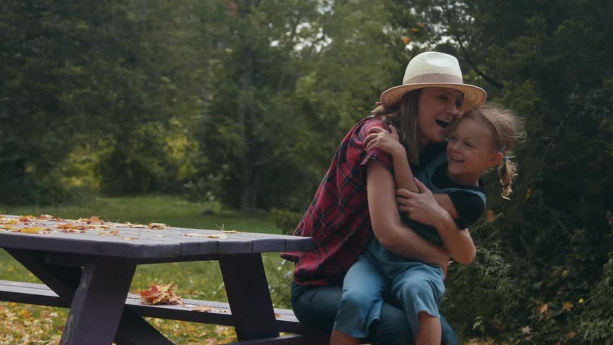 A little girl runs over to her mother who is sitting at a grey park bench and gives her a hug during the daytime