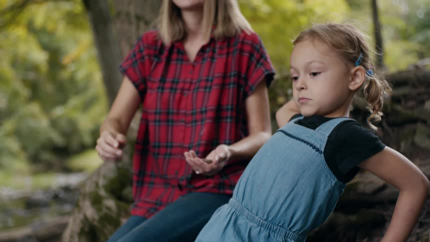 A mother hands her daughter a small rock to throw while they sit in the forest