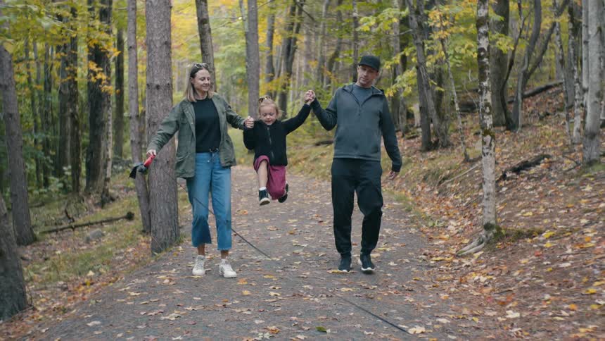 A mom and dad swings their daughter by their hands as they walk through a forest on a paved path during the day