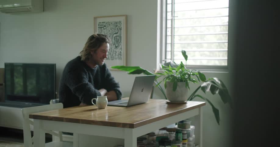 A man sits at a table having a video call on a laptop and drinks from a small mug