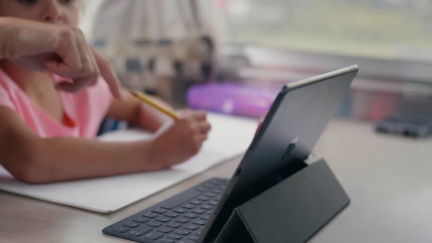 Young girl sits with pencil in hand looking at what an adult is pointing to on a tablet