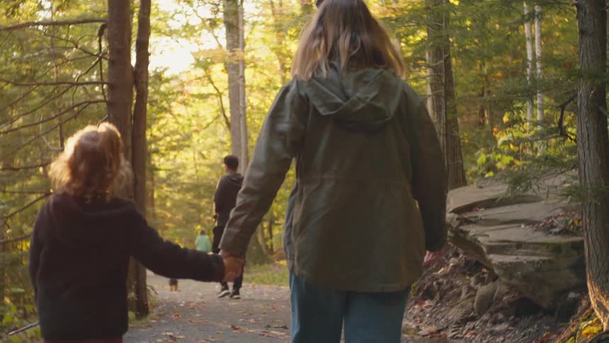 A mom and daughter hold hands as they walk on a paved path in a forest during the day