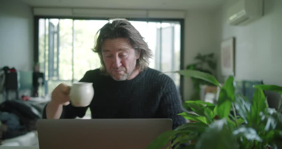 Man with long hair and a black pullover makes a video call on his laptop while drinking a cup of coffee